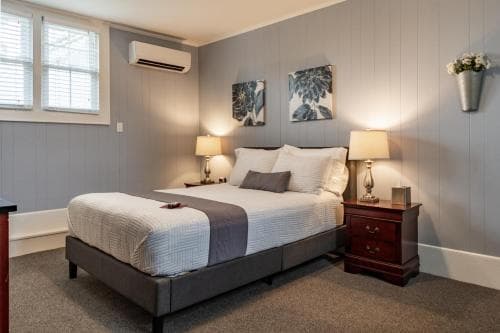 A cozy hotel room features a neatly made bed with plush pillows, warm lighting from bedside lamps, framed botanical artwork on gray walls, and a window with natural light, ideal for travelers seeking comfort and relaxation.