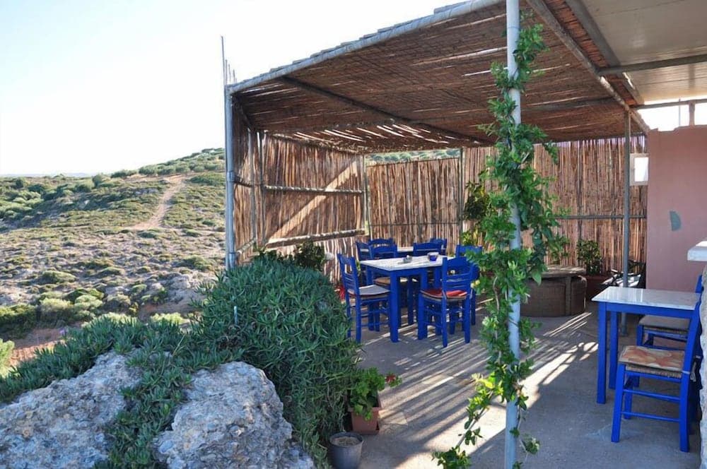 A serene outdoor dining area with blue chairs and tables under a thatched roof, surrounded by lush greenery and rocky terrain, offers a tranquil retreat ideal for travelers seeking relaxation in nature.