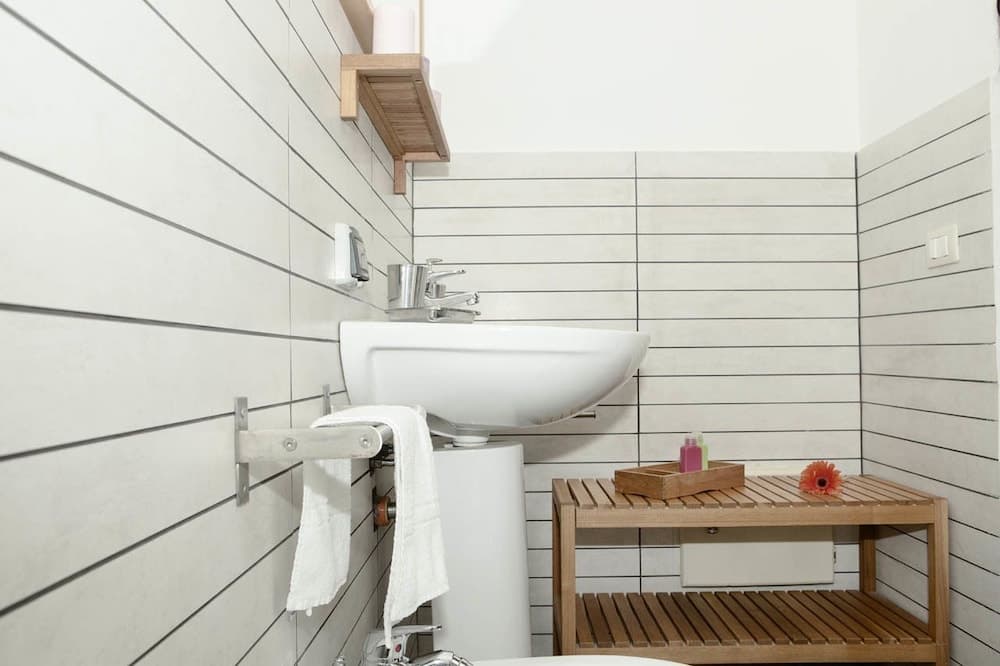 A serene minimalist bathroom features sleek white tiles, a modern sink, wooden shelving with toiletries, and a fresh orange flower, ideal for travelers seeking a clean and tranquil accommodation space.