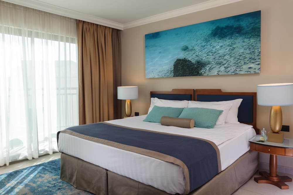 A serene hotel room features a cozy bed adorned with teal pillows, a large aquatic artwork on the wall, and a balcony view through sheer curtains, ideal for travelers seeking relaxation and comfort.
