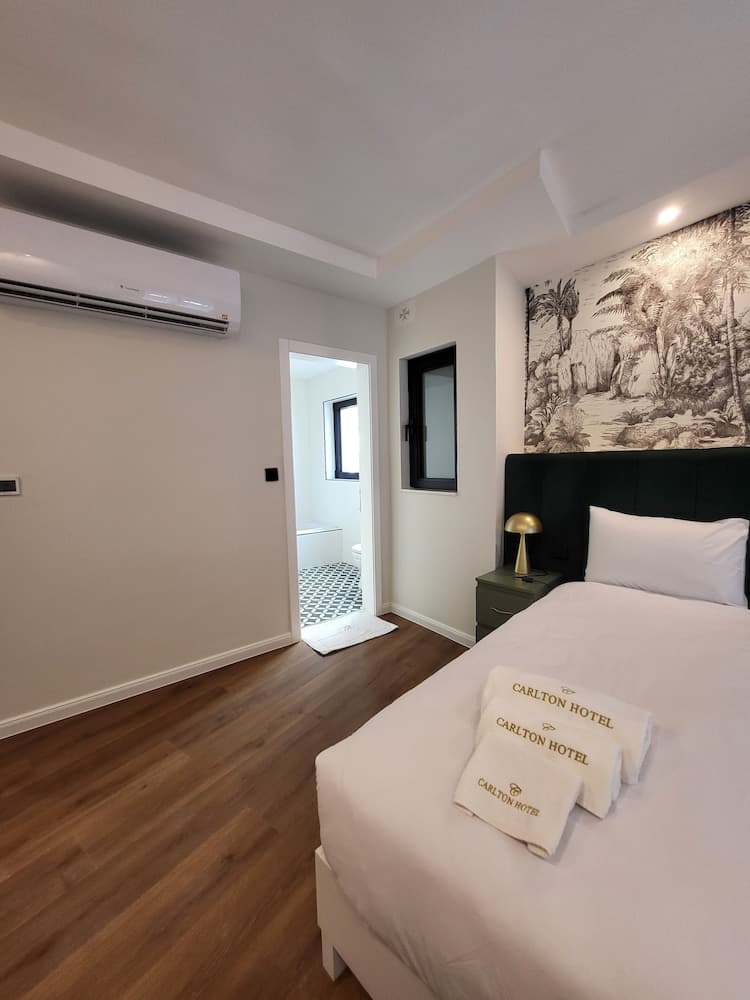 A cozy hotel room features a neatly made bed with Carlton Hotel towels, a tropical mural wall, wooden flooring, and an en-suite bathroom, ideal for travelers seeking comfort and style.