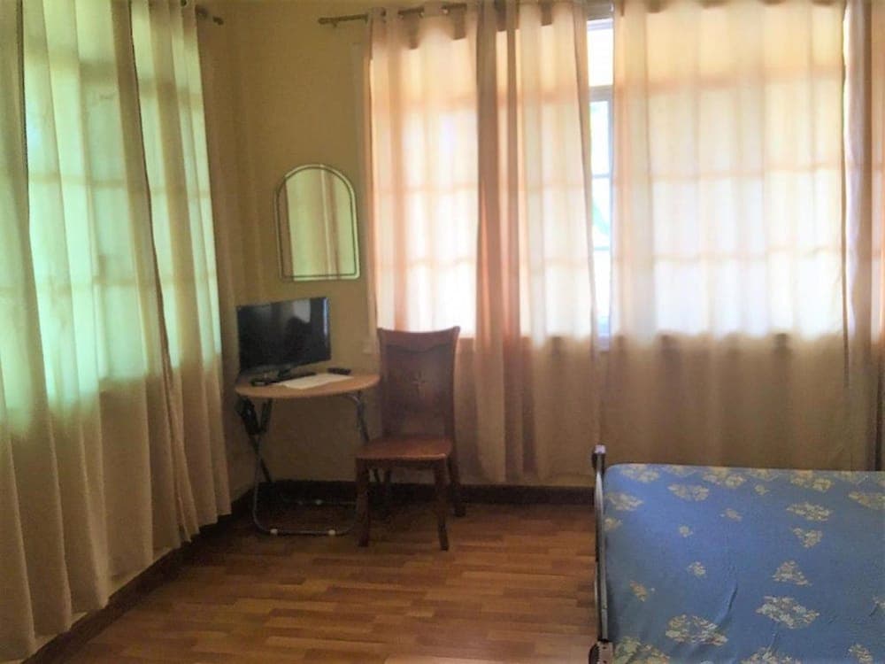 A cozy hotel room with wooden floors, sheer curtains, a small desk with a TV, and a floral bedspread offers a comfortable and simple stay for budget travelers.