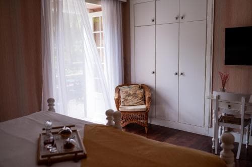 A cozy hotel room features a neatly made bed, a wicker chair by the window with sheer curtains, and a small dining table, creating a serene and inviting atmosphere ideal for travelers seeking relaxation.