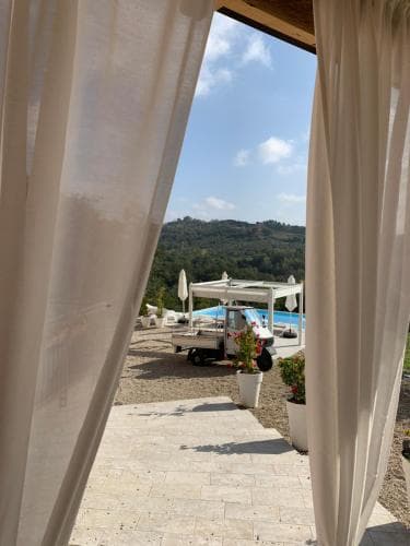 A serene outdoor terrace framed by flowing curtains offers a picturesque view of lush hills, a vibrant poolside area with umbrellas, and potted plants, ideal for a relaxing vacation getaway.