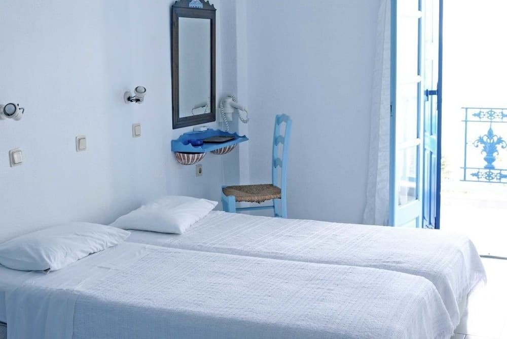 A serene Mediterranean-style bedroom with white walls, twin beds, a blue chair, and an open door leading to a balcony, ideal for travelers seeking a tranquil coastal retreat.