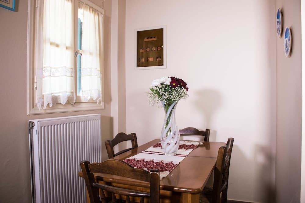 A cozy dining nook features a wooden table adorned with a floral centerpiece, set against soft beige walls with decorative plates and a window framed by lace curtains, ideal for travelers seeking a quaint and inviting accommodation.