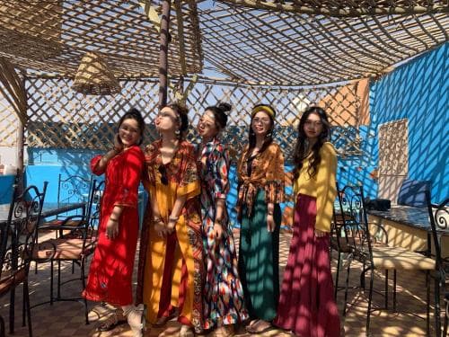 Five women in vibrant dresses stand under a rustic wooden pergola casting dappled shadows, creating a cheerful and relaxed atmosphere ideal for social gatherings or cultural exploration in an outdoor café setting.