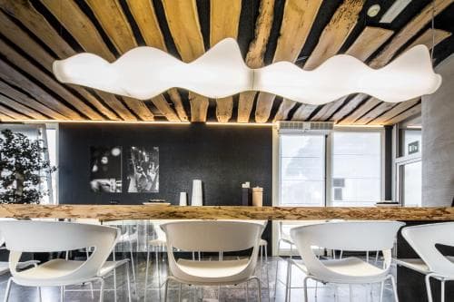 A modern dining area features sleek white chairs around a long wooden table under a wavy white light fixture against a rustic wooden ceiling, ideal for travelers seeking contemporary design in urban accommodations.