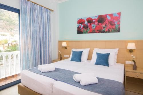 A cozy hotel room with a neatly made bed adorned with blue accents, a vibrant floral artwork on the wall, and a balcony offering a serene view of lush greenery, ideal for travelers seeking relaxation and comfort.