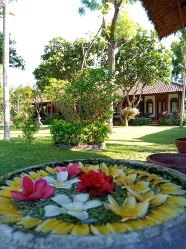 A serene tropical garden features vibrant floral arrangements in a stone basin, surrounded by lush greenery and traditional thatched-roof bungalows, ideal for a tranquil vacation retreat.