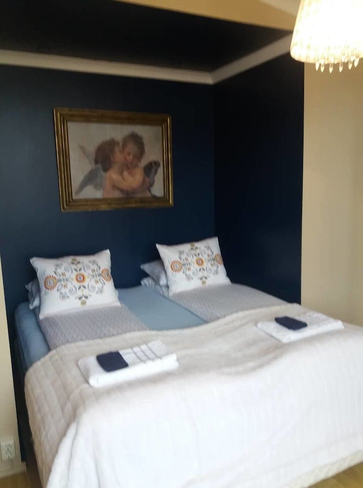 A cozy hotel room features two neatly arranged beds with patterned pillows against a dark blue wall adorned with an angelic artwork, illuminated by a warm chandelier, ideal for travelers seeking comfort and tranquility.