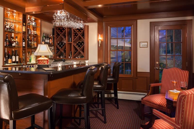 A cozy bar area at the Harbor Light Inn in Marblehead, MA, features leather stools, a well-stocked liquor cabinet, warm lighting, and inviting seating by large windows, perfect for travelers seeking a relaxing evening ambiance.