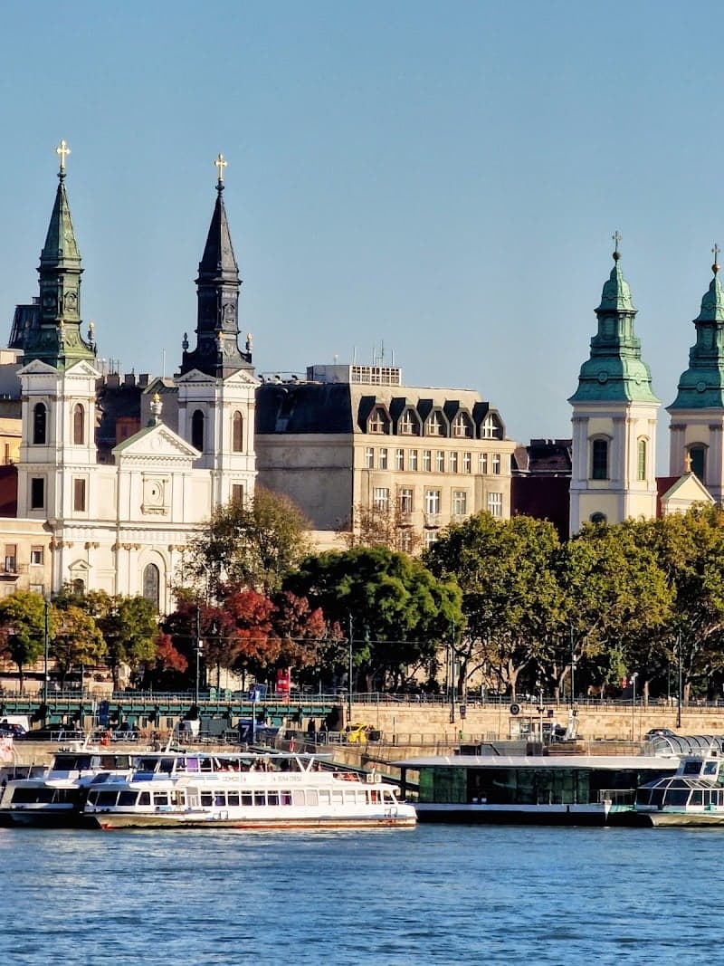 The serene Inner City of Budapest showcases historic churches with spires against a clear sky, framed by lush trees and riverboats along the Danube, ideal for cultural exploration and scenic river cruises.