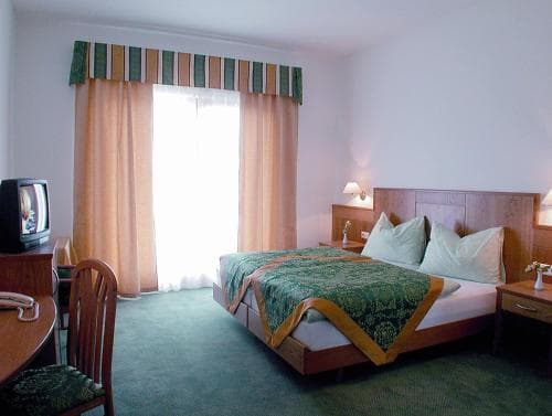 A cozy hotel room features a neatly made bed with green bedding, warm-toned curtains framing a bright window, a small dining area, and a vintage TV, ideal for travelers seeking comfort and simplicity.