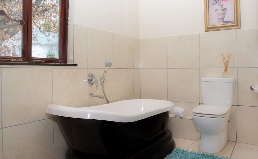 A cozy bathroom with beige tiled walls features a stylish black clawfoot bathtub, a white toilet adorned with a reed diffuser, and a window offering a serene outdoor view, ideal for travelers seeking relaxation and comfort.