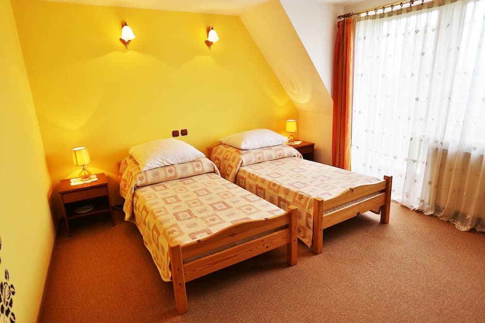 A cozy hotel room with two wooden twin beds, yellow walls, soft lighting, and sheer curtains creates a warm and inviting atmosphere ideal for travelers seeking comfort and relaxation.