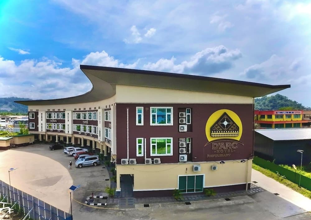 A modern hotel with a distinctive curved roof and cultural emblem stands invitingly under a bright sky, surrounded by parked cars and lush greenery, ideal for travelers seeking comfort and local charm.