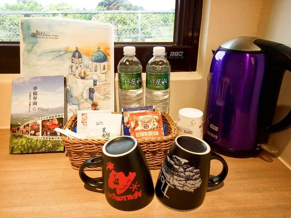 A cozy hotel room setup features a wooden table with travel brochures, bottled water, a basket of snacks, two decorative mugs, and a purple electric kettle, creating a welcoming atmosphere for travelers seeking refreshments.