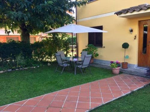 A cozy backyard patio with a tiled pathway, lush greenery, and a white umbrella over seating invites travelers seeking a tranquil retreat in a quaint residential area.