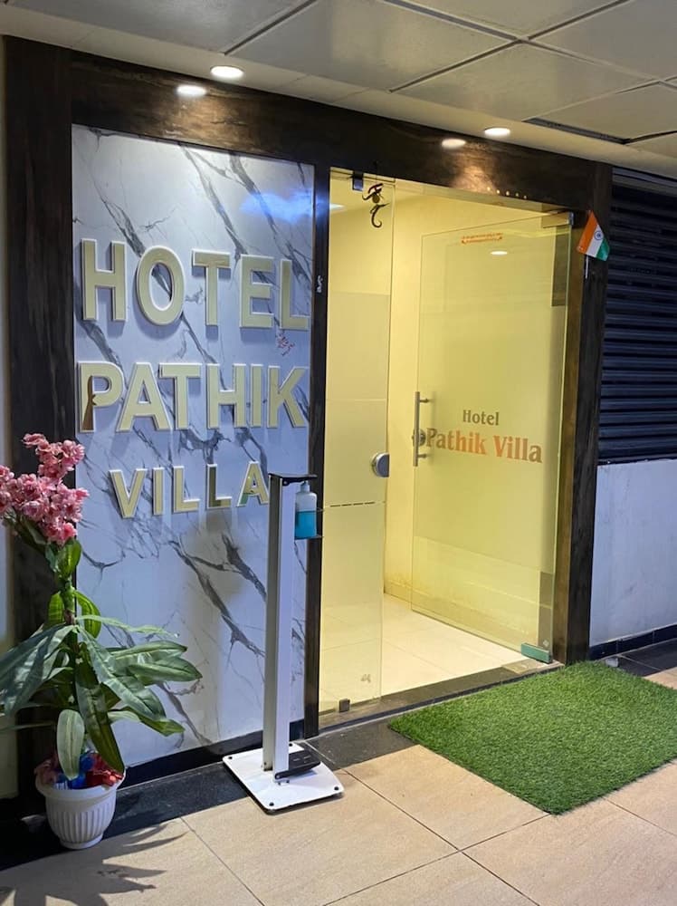 A welcoming entrance to Hotel Pathik Villa features a modern glass door, a marble signboard, a potted plant with pink flowers, and a hand sanitizer stand, ideal for travelers seeking a clean and inviting accommodation.