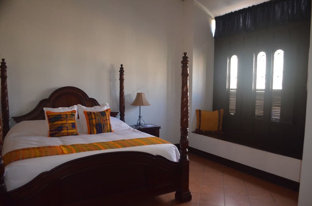 A cozy bedroom with a four-poster bed adorned in vibrant plaid textiles, set against white walls and illuminated by natural light through arched window shutters, offers a serene retreat ideal for travelers seeking comfort and charm.