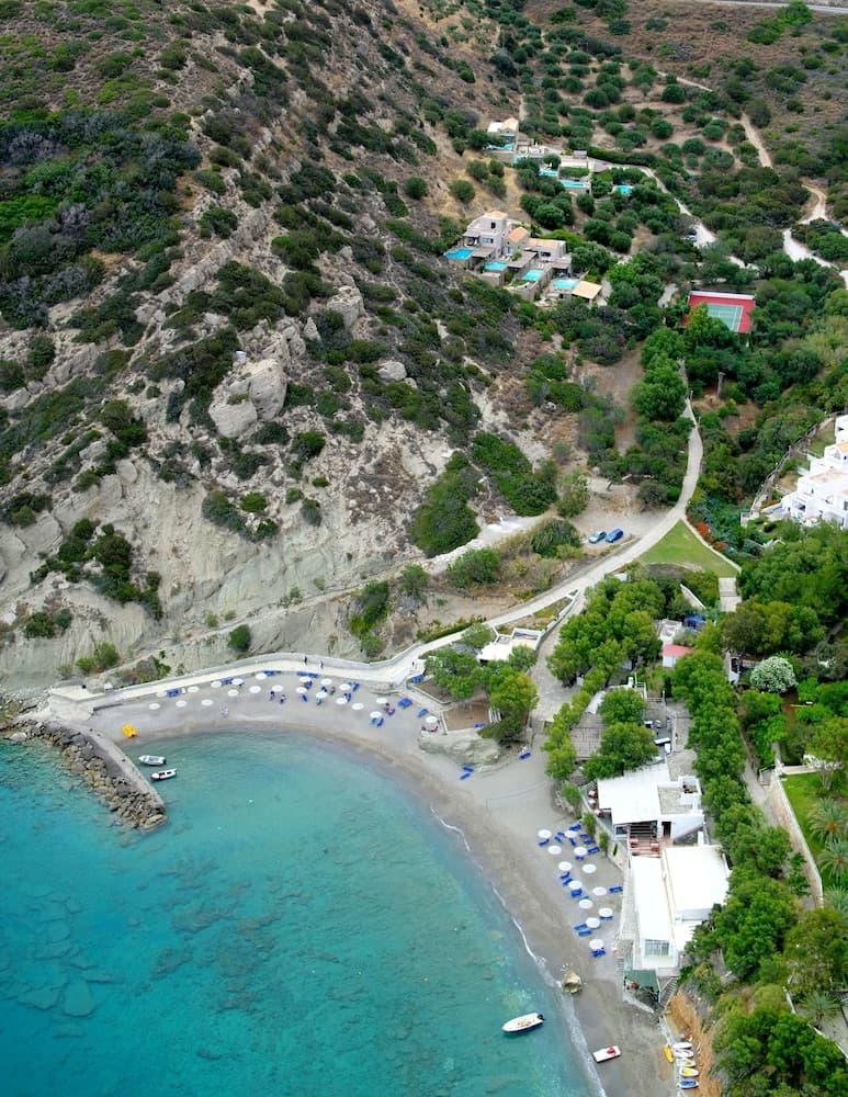An aerial view captures a serene coastal village with turquoise waters, sandy beaches lined with umbrellas, lush greenery, and scattered villas, ideal for a tranquil Mediterranean getaway.
