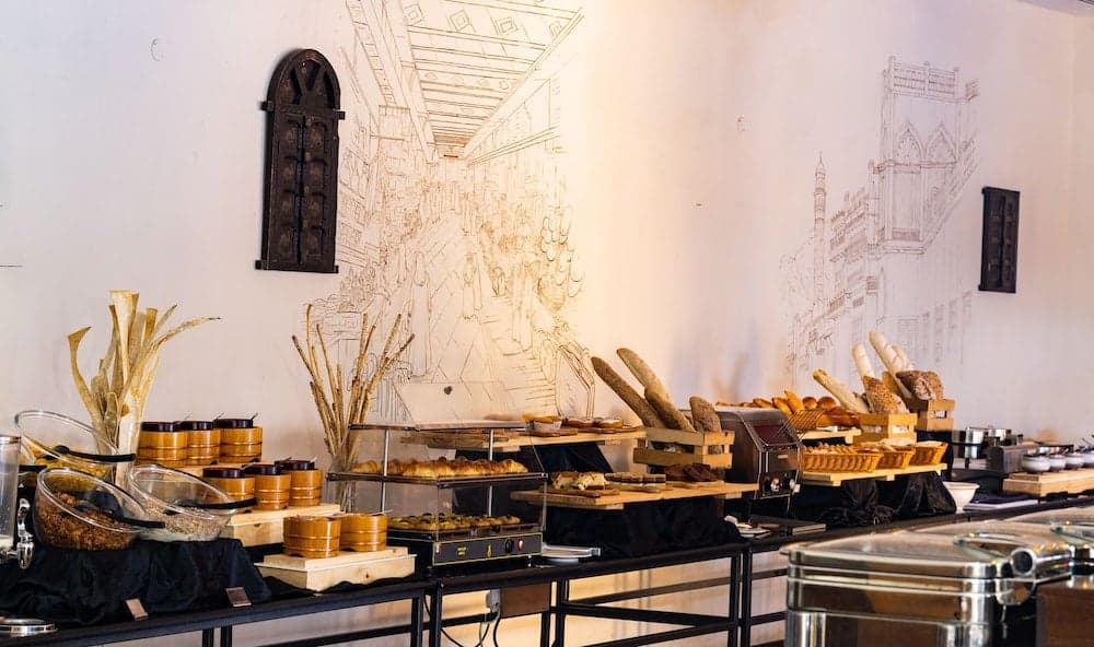 A cozy café buffet scene features an array of breads, pastries, and cereals under warm lighting, set against a wall adorned with architectural sketches, ideal for travelers seeking a charming breakfast spot.