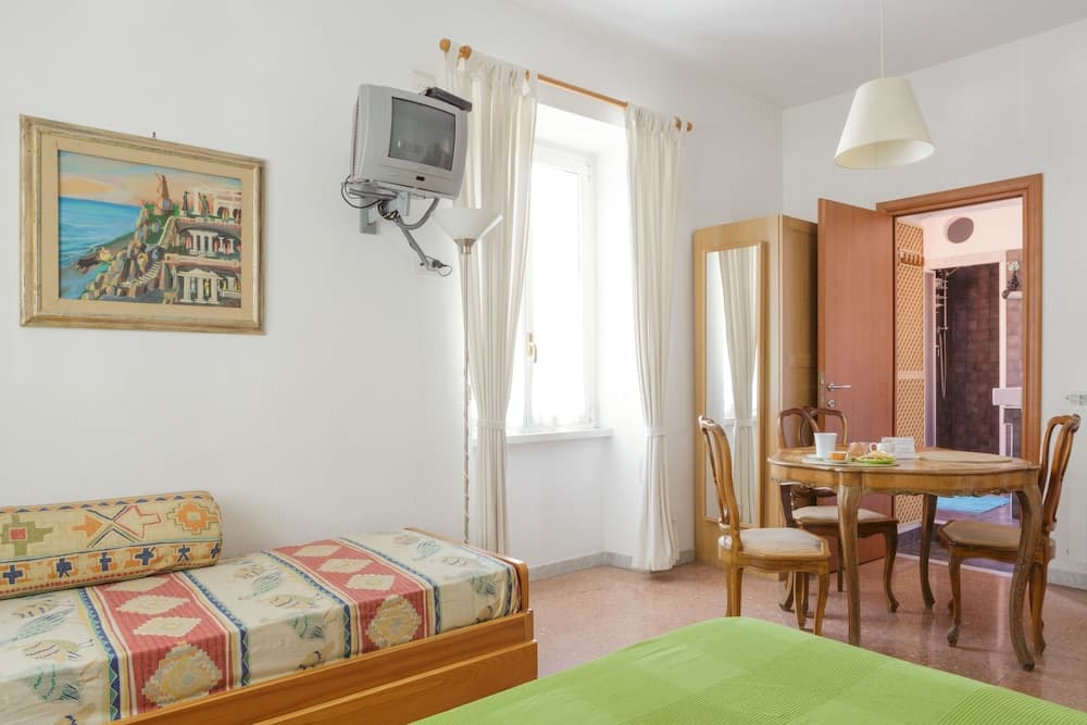 A cozy hotel room features twin beds with colorful linens, a wooden dining set by the window, and a wall-mounted TV, ideal for travelers seeking comfort and simplicity.