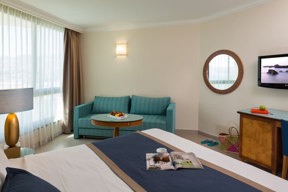 A cozy hotel room features a neatly made bed, turquoise sofa, round mirror, and large window with sheer curtains, creating a serene and inviting atmosphere ideal for travelers seeking relaxation.