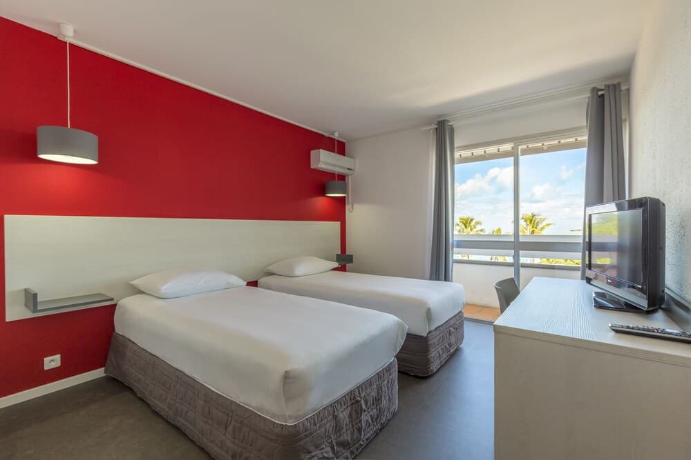 A cozy hotel room with two neatly made beds, a vibrant red accent wall, and a balcony view of palm trees and the ocean, ideal for travelers seeking comfort and a tropical escape.