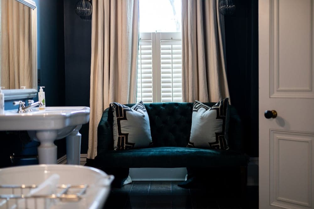 A cozy bathroom corner features a plush teal velvet sofa adorned with patterned pillows, set against dark walls with soft curtains and shutters, creating an inviting and luxurious retreat ideal for boutique hotel stays.