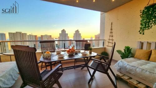 A cozy balcony setup with wooden furniture and a vibrant fruit centerpiece overlooks a stunning cityscape at sunset, ideal for urban travelers seeking relaxation and scenic views.