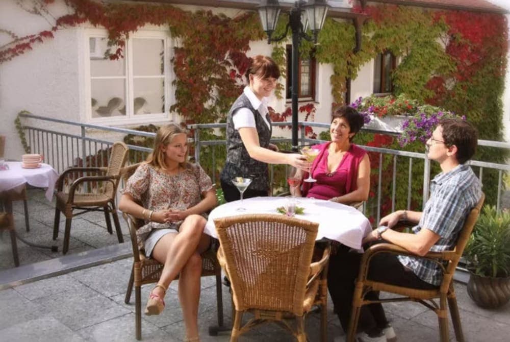 A cozy outdoor terrace adorned with autumnal vines and flowers sets the scene for a relaxed dining experience, where a waitress serves drinks to guests seated at wicker chairs around a white-clothed table, ideal for travelers seeking a charming and intimate restaurant setting.