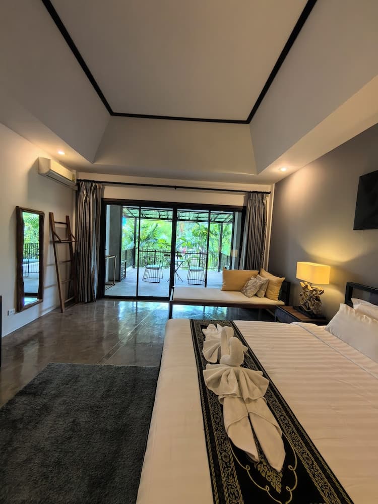 A serene hotel bedroom with a neatly made bed adorned with decorative towels, a cozy seating area by the glass doors leading to a lush balcony, and modern amenities, perfect for a relaxing tropical getaway.