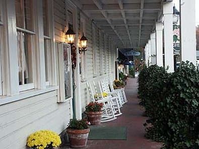 A charming covered porch lined with white rocking chairs, potted flowers, and vintage lanterns creates a cozy, inviting atmosphere perfect for a leisurely getaway at a quaint bed and breakfast.