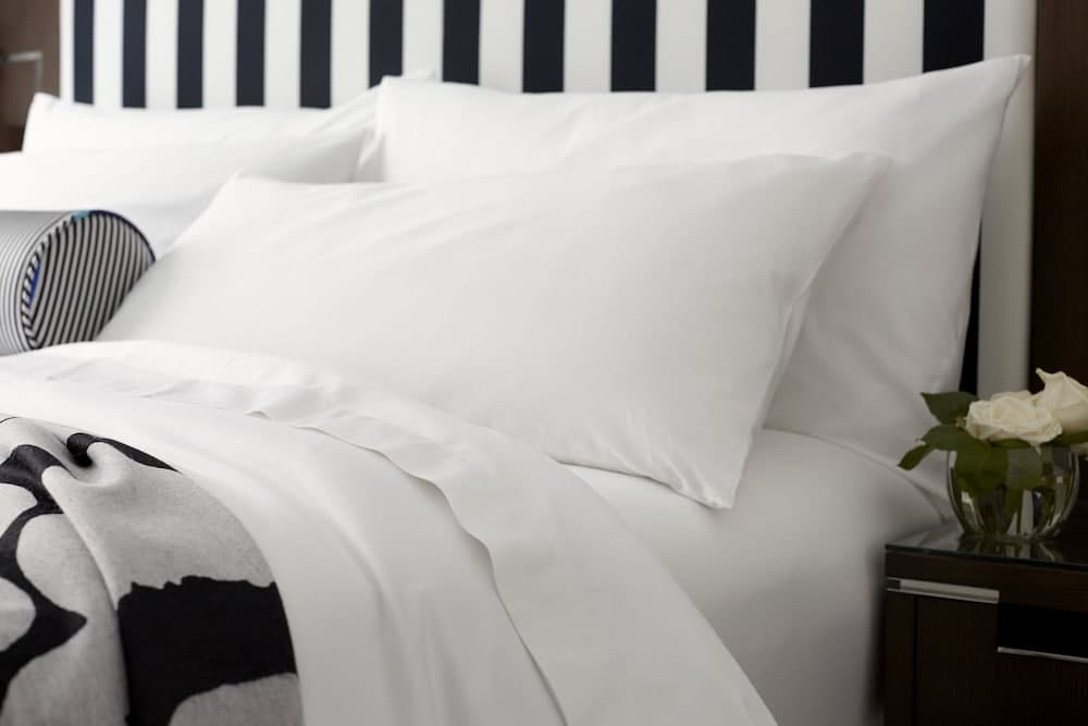A serene hotel bedroom scene features crisp white linens on a bed with a black-and-white striped headboard, complemented by a vase of white roses on the bedside table, ideal for travelers seeking comfort and style.
