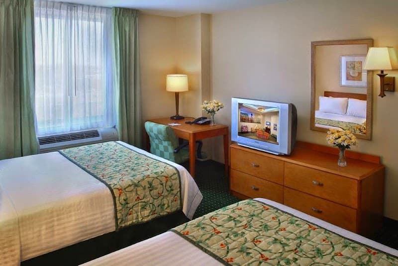 A cozy hotel room in Corona, NY features two neatly made beds with floral quilts, a wooden desk with a lamp, a TV on a dresser adorned with flowers, and a window with sheer curtains, ideal for travelers seeking comfort and convenience.