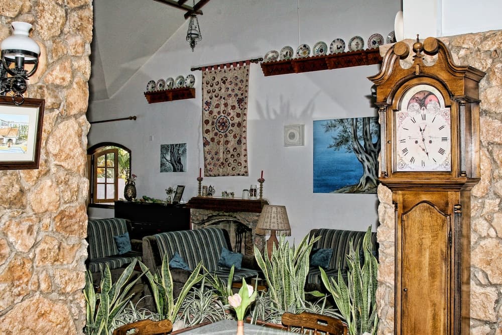 A cozy living room with stone walls, striped sofas, lush indoor plants, and a wooden grandfather clock creates a warm and inviting atmosphere, perfect for travelers seeking a charming and rustic retreat.