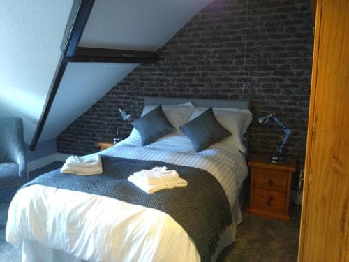 A cozy attic bedroom with exposed brick walls, a neatly made bed adorned with gray pillows, and wooden furnishings creates an inviting atmosphere perfect for travelers seeking a charming and intimate accommodation experience.