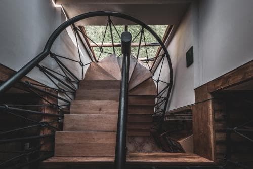 A modern spiral staircase with wooden steps and geometric metal railings ascends toward a bright window, creating an inviting and stylish atmosphere ideal for exploring urban loft spaces.