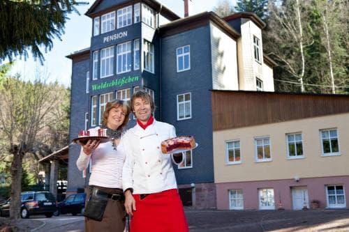 A cheerful couple presents baked goods in front of a charming pension building nestled among trees, ideal for travelers seeking a cozy retreat with homemade treats.