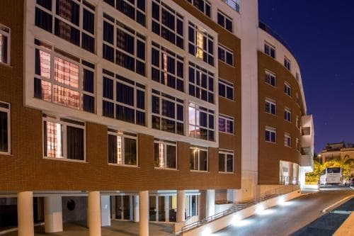 A modern multi-story building illuminated at night with warm lights through its windows creates an inviting urban atmosphere, ideal for travelers seeking accommodation in a bustling city center.
