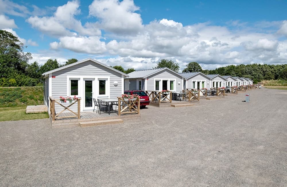 A serene row of cozy cottages with wooden decks and vibrant flower boxes lines a gravel path under a partly cloudy sky, ideal for a tranquil family vacation retreat.