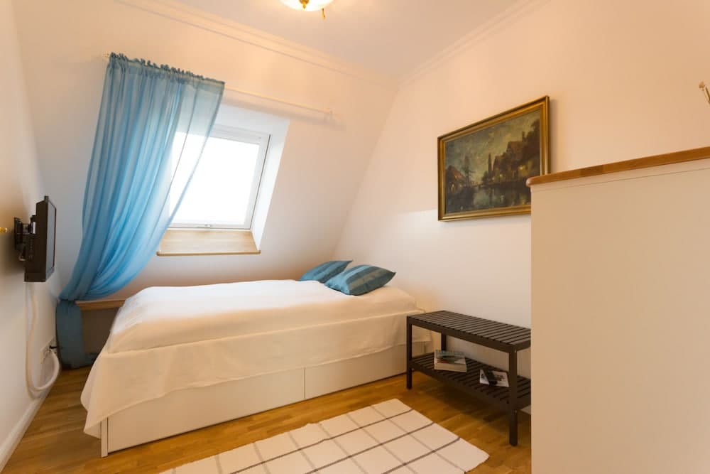A cozy attic bedroom with a single bed, blue curtains, wooden flooring, and a framed painting creates a serene atmosphere ideal for travelers seeking a quaint and comfortable stay.