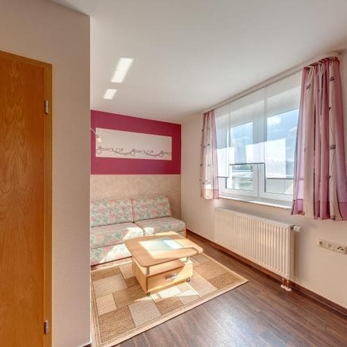 A cozy apartment living room with wooden flooring, a floral-patterned sofa, pink curtains, and ample natural light through a large window, ideal for travelers seeking a comfortable and homey stay.