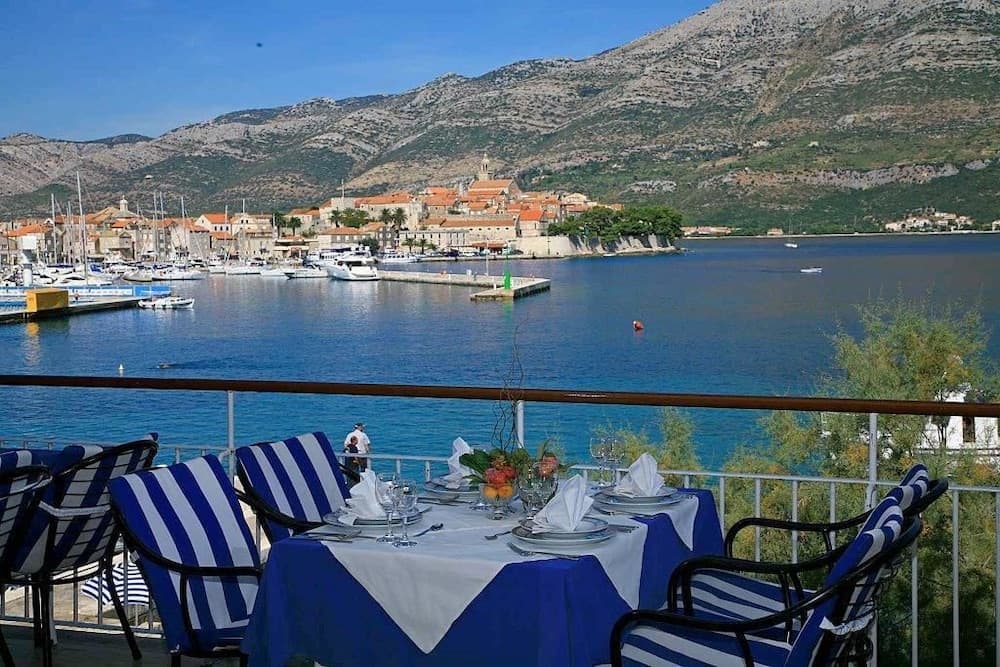 A serene coastal dining terrace overlooks a picturesque harbor with docked boats and a charming town nestled against rugged mountains, ideal for travelers seeking a tranquil seaside retreat.