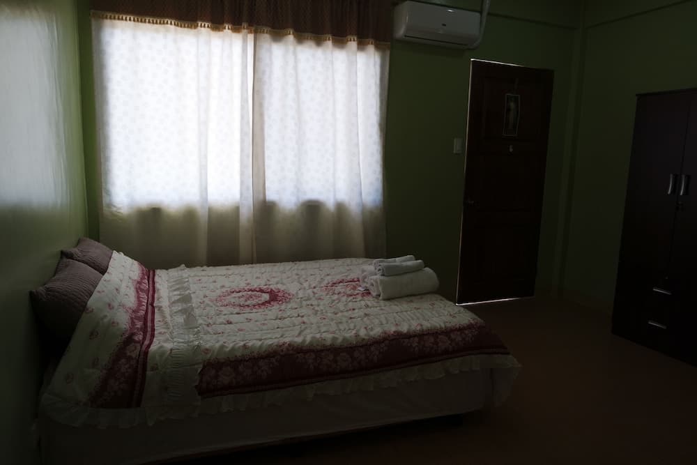 A cozy bedroom with green walls, a neatly made bed adorned with a floral quilt, soft curtains filtering natural light, and essential furnishings creates a serene atmosphere ideal for travelers seeking a peaceful retreat.