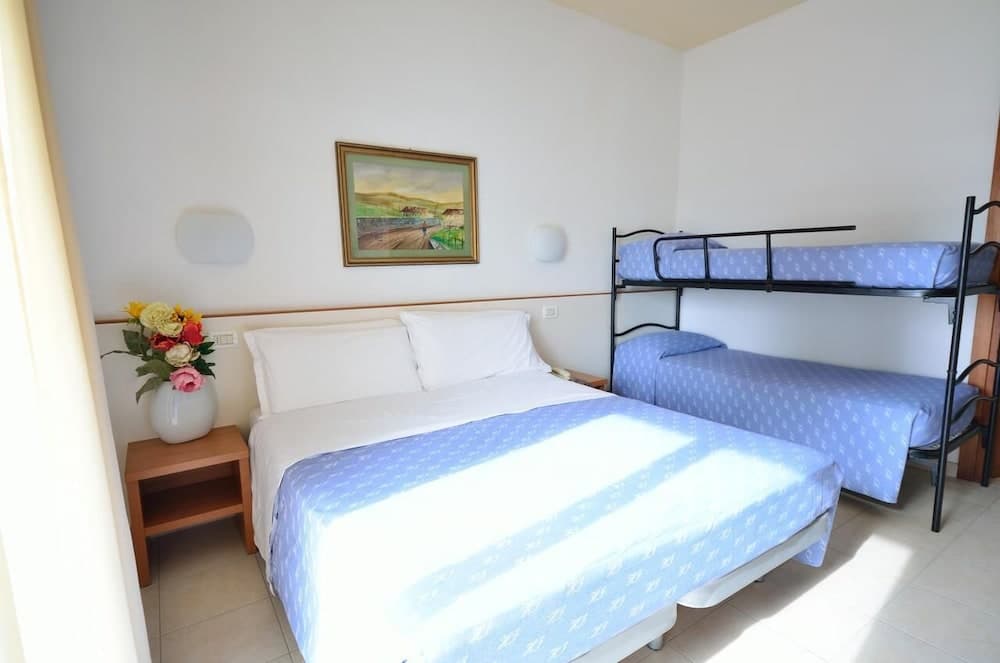 A cozy hotel room features a neatly made double bed beside a bunk bed, adorned with blue linens, a floral vase on a wooden nightstand, and a serene landscape painting, ideal for family travelers seeking comfort and simplicity.