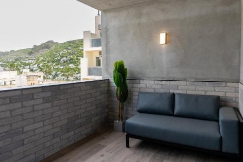 A cozy balcony with a modern blue sofa and a tall potted plant offers a serene view of lush green hills and white buildings, ideal for travelers seeking relaxation in a contemporary urban retreat.