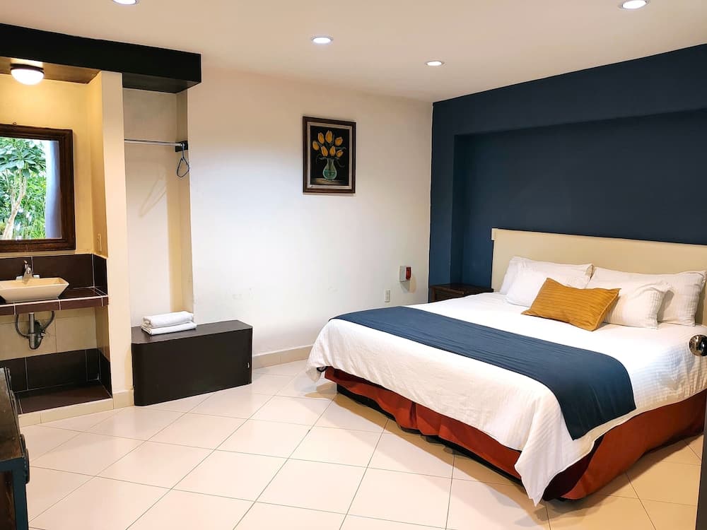 A cozy hotel room with a neatly made bed, modern bathroom sink area, and a framed floral artwork creates a serene atmosphere ideal for travelers seeking comfort and relaxation.